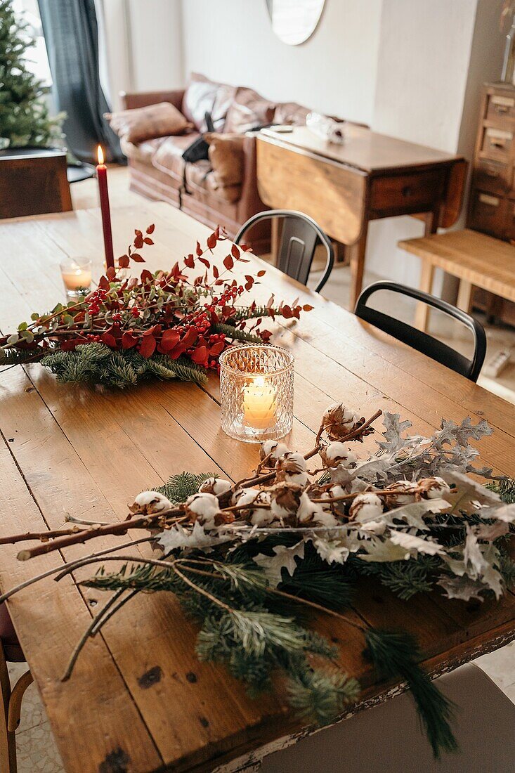 From above of festive Christmas bouquet with branches of cotton, fir and twigs of eucalyptus and bright red branches with berries placed on wooden table with candles in room