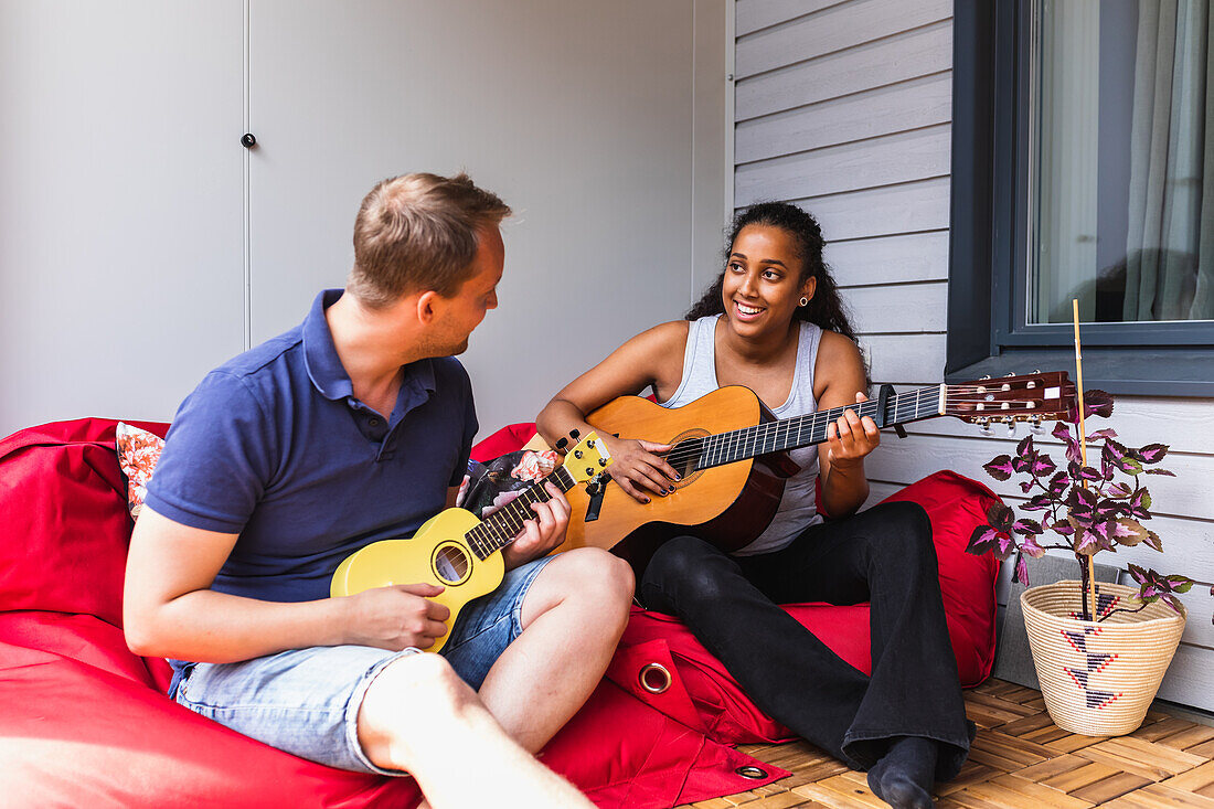 Junges, fröhliches, multiethnisches Paar sitzt auf Sitzsäcken auf der gemütlichen, hellen Terrasse einer Wohnung und spielt Gitarre und Ukulele und sieht sich dabei an