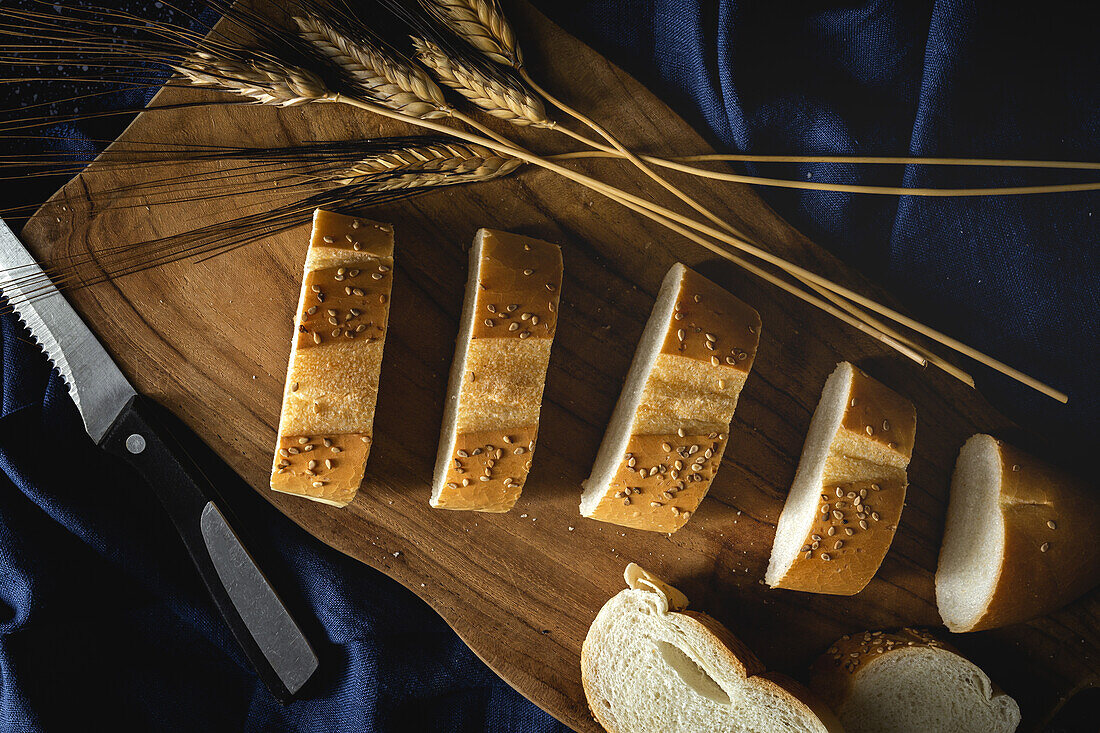 Top view of pieces of white bread near knife and wheat spikes on wooden board