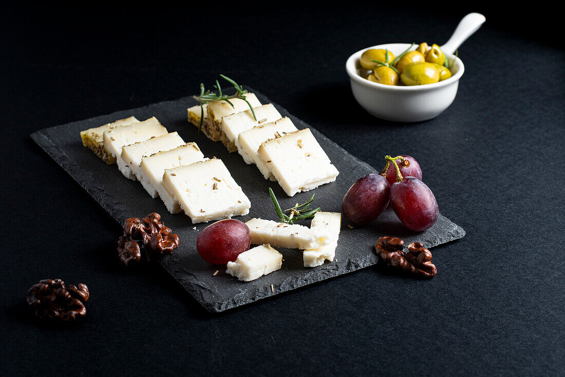 Leckere frische Käsescheiben und reife Weintrauben auf einem schwarzen Brett mit Nüssen und Rosmarinzweigen