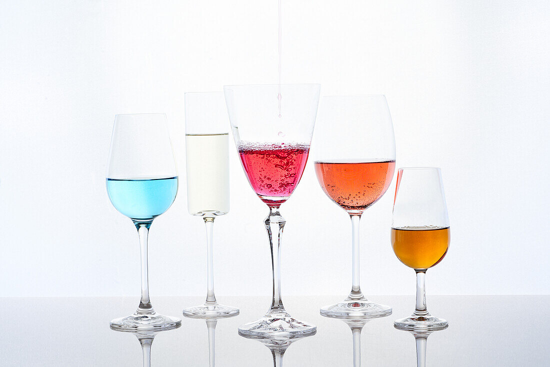 Assorted colorful cocktails in various shaped crystal glasses served on mirrored table against white background