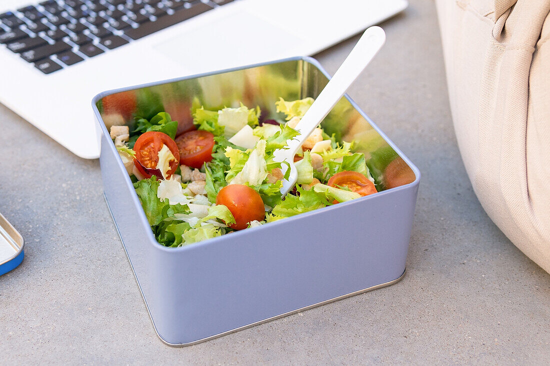 Salad with fresh green lettuce and cherry tomatoes served in square metal container with plastic spoon placed near modern netbook