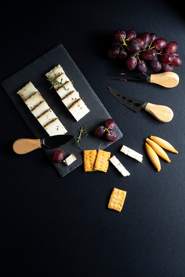 Top view of assorted delicious cheese arranged on black board with ripe grapes and crackers placed near knifes on dark surface