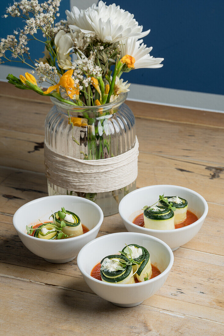 Von oben appetitliche Zucchiniröllchen in weißen Schalen mit Tomatensoße neben Glasvase mit frischem blühendem Strauß auf Holzboden serviert