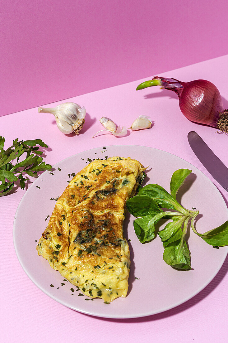 Tasty omelette on plate against fresh parsley sprigs and red onion with garlic cloves on pink background