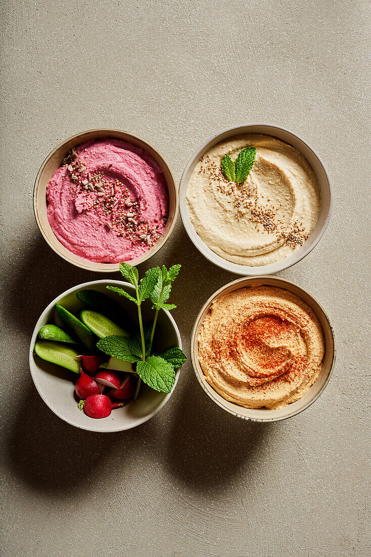 Top view of bowls with assorted hummus served on table with fresh cucumbers and radish