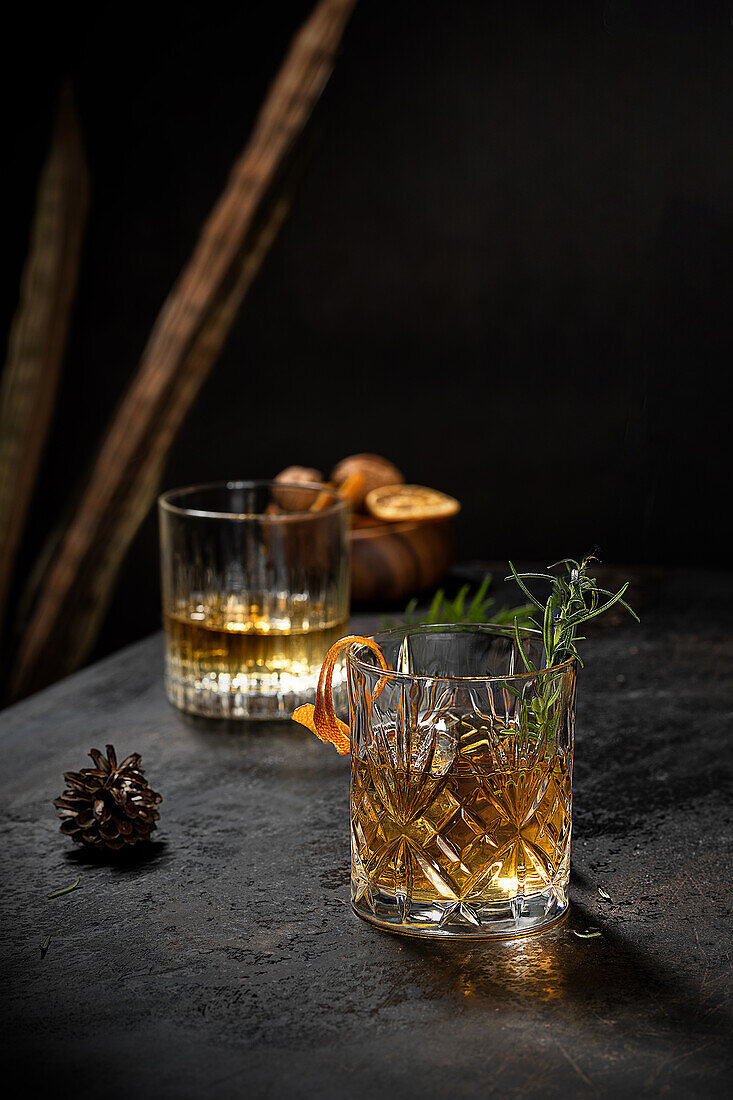 Kristallglas mit altmodischem Whiskey, garniert mit frischem Rosmarin und Orangenschalen auf einem schwarzen Tisch