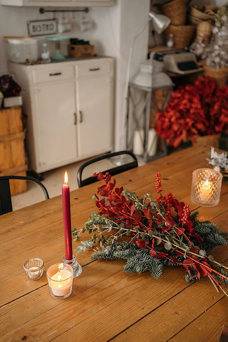 Festlicher, stilvoller, dekorativer Weihnachtsstrauß mit Eukalyptuszweigen und leuchtend roten Zweigen mit Beeren auf einem Holztisch mit Kerzen im Raum von oben