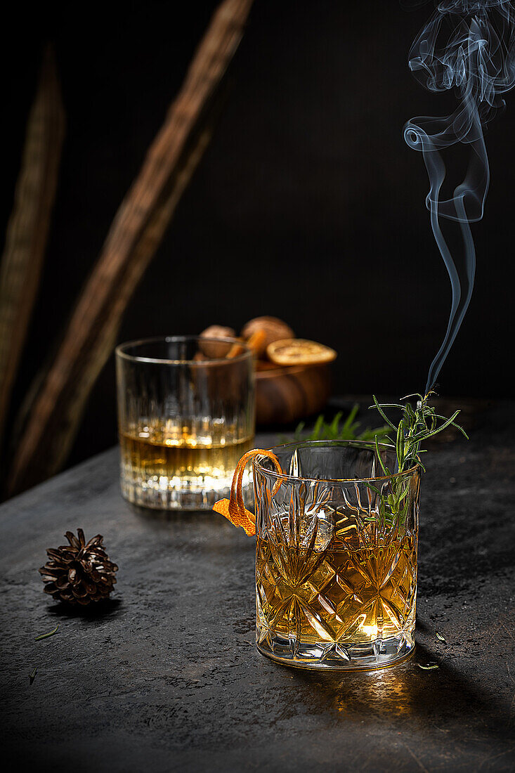 Crystal glass of old fashioned whiskey drink garnished with fresh rosemary and orange peel on black table