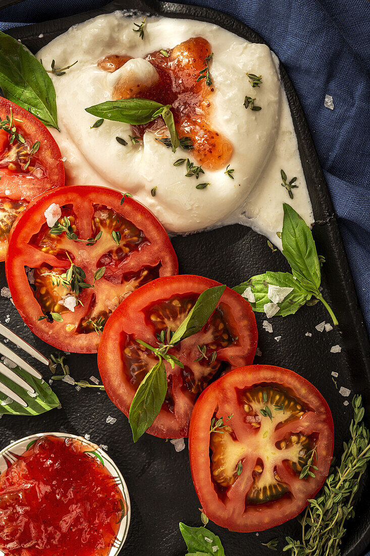 From above of appetizing burrata cheese with sauce and slices of fresh tomato with thyme and basil leaves served on cast iron tray placed on table cloth on wooden table