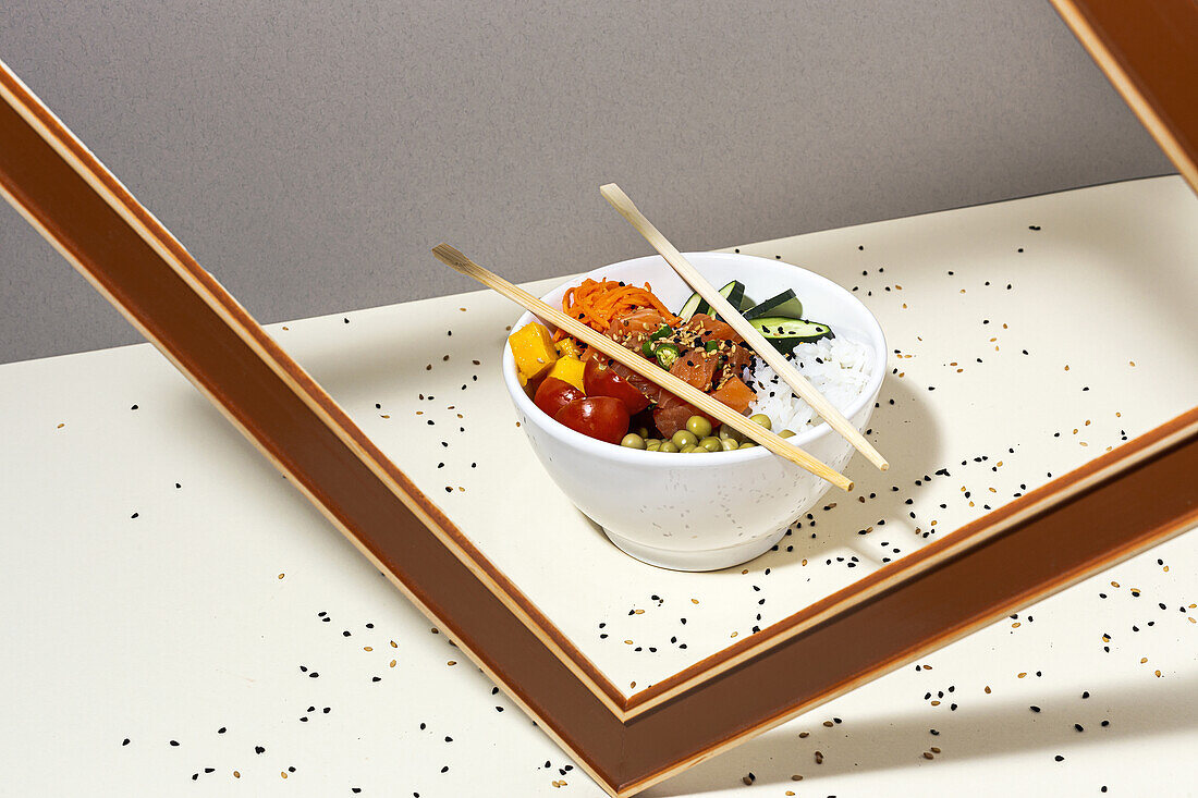 Weiße Schale mit schmackhaftem Poke-Gericht und Essstäbchen hinter dem Rahmen auf einem mit Sesam bedeckten Tisch