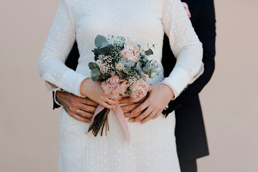 Anonymer Bräutigam umarmt elegante Braut in weißem Hochzeitskleid mit zartem Blumenstrauß