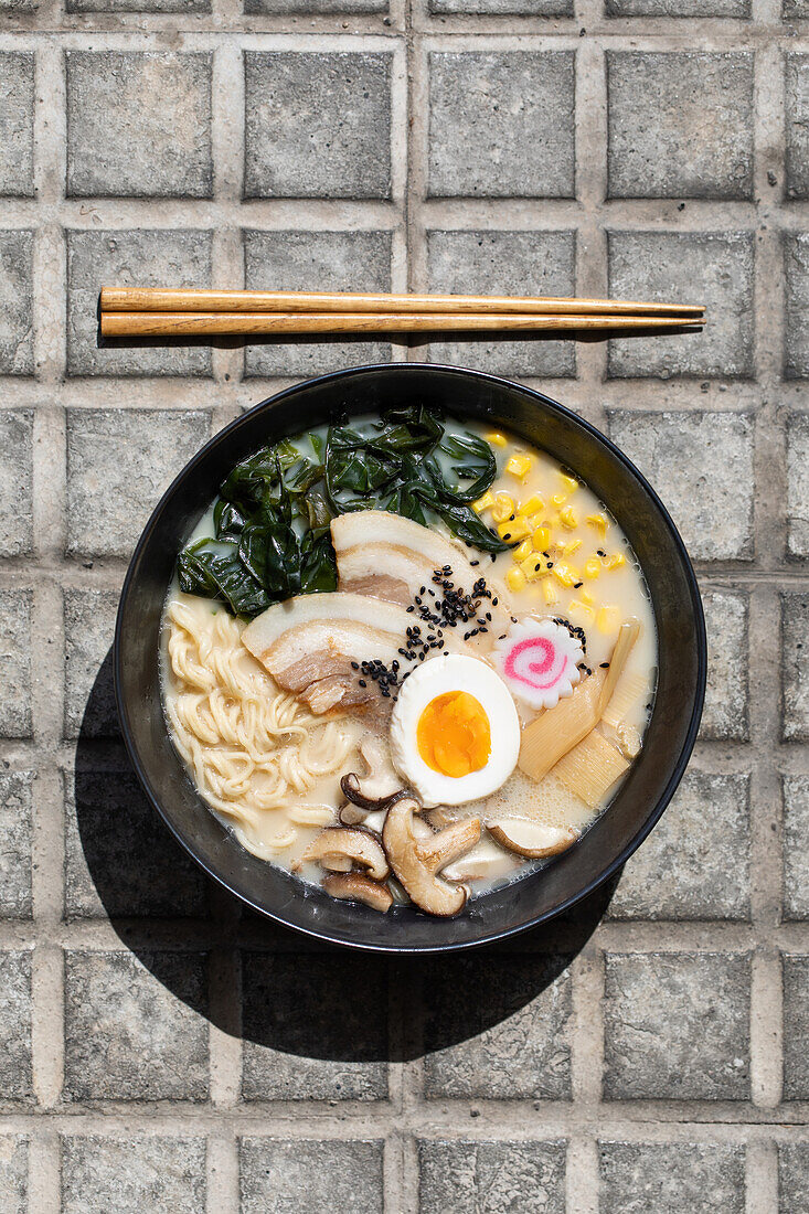 Draufsicht auf appetitliche japanische Ramen mit Pilzen und Ei, serviert auf dem gepflasterten Bürgersteig mit Stäbchen in einer sonnigen Straße in der Stadt