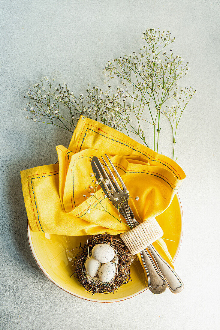 From above place setting for Easter dinner with yellow ceramic plates near nest with easter eggs surrounded by cushion baby's-breath leaves