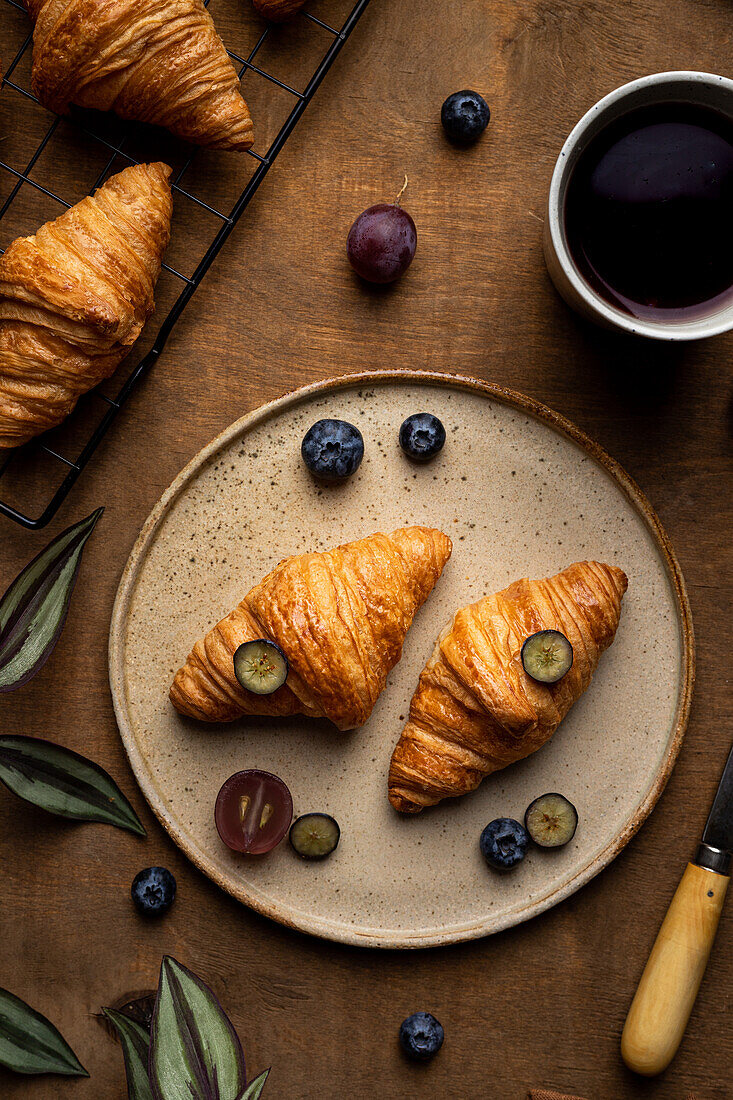 From above of tasty fresh baked croissants served on plate with fruits placed near cup of tea on wooden table in morning time in light room