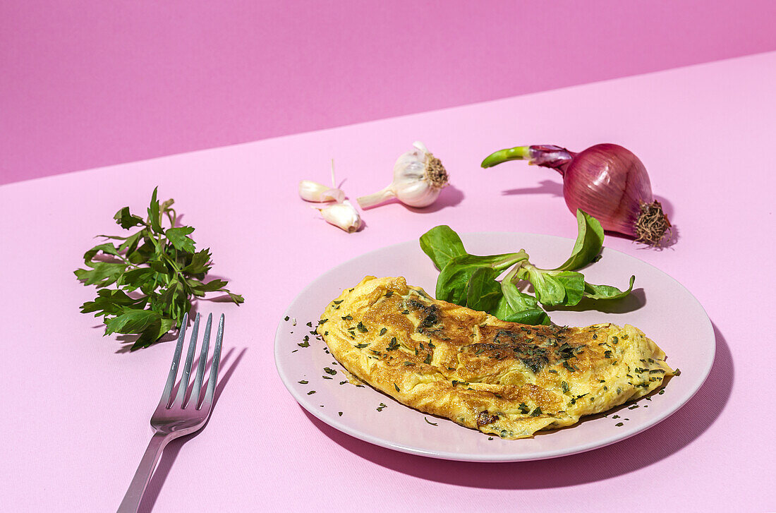 Tasty omelette on plate against fresh parsley sprigs and red onion with garlic cloves on pink background