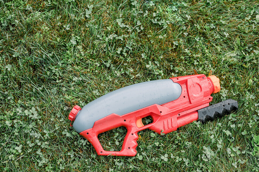From above of plastic red and gray toy water pistol placed on grassy lawn in park