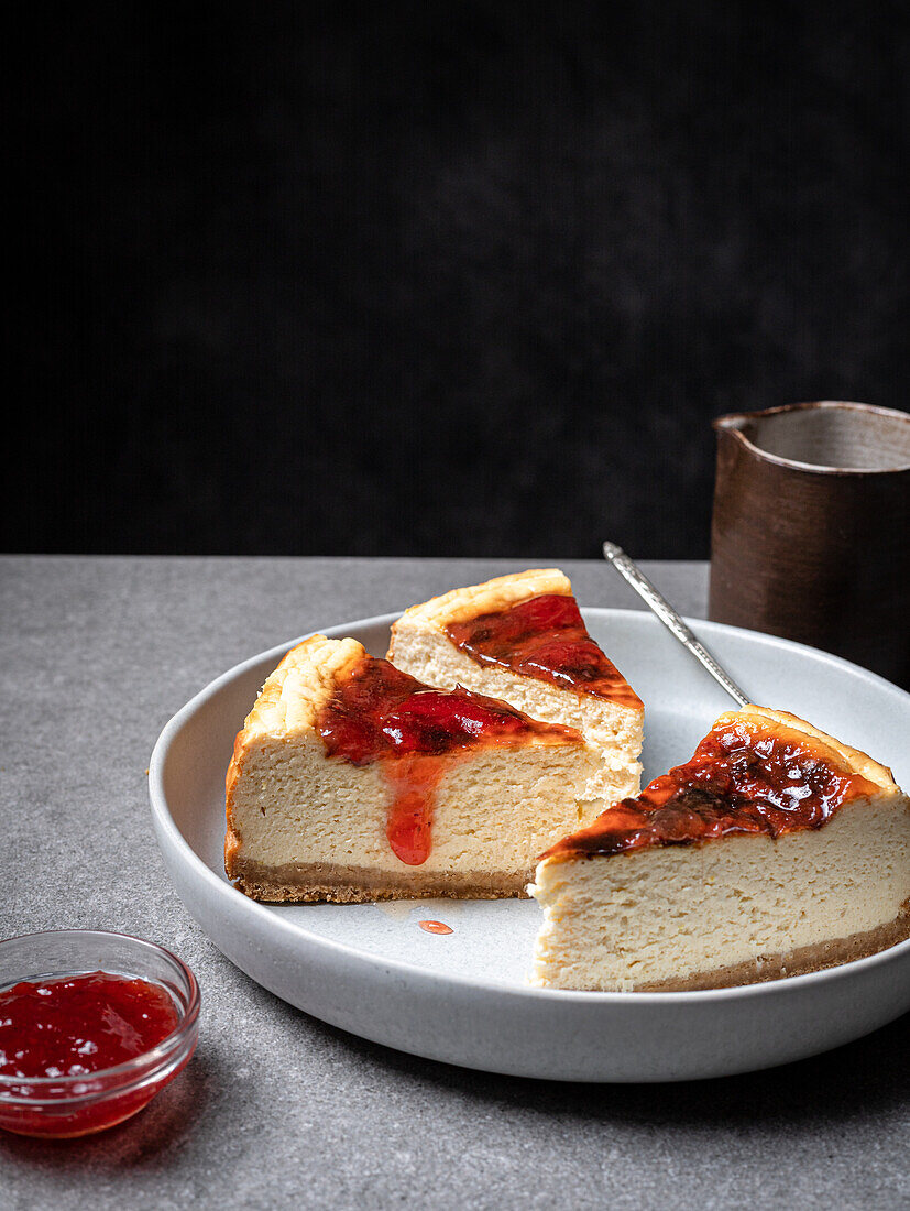 Teller mit in Scheiben geschnittenem köstlichem hausgemachtem Käsekuchen und Glas mit süßer Marmelade auf grauem Tisch