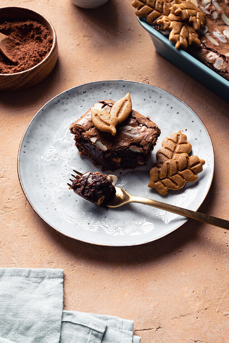 Ein Stück süßer, frisch gebackener Schokoladen-Brownie mit Keksen, serviert auf einem Teller neben einer Backform auf einem Tisch mit einer Tasse Kaffee