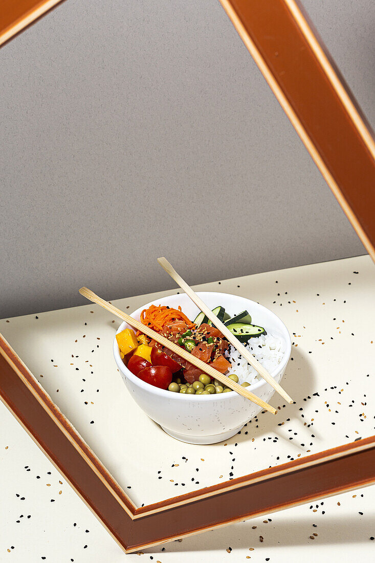 High angle of white bowl with tasty poke dish and chopsticks placed behind frame on table covered with sesame seeds