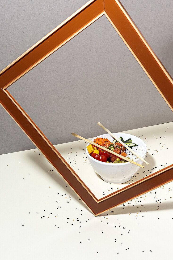 Hoher Winkel einer weißen Schale mit leckerem Poke-Gericht und Stäbchen, die hinter dem Rahmen auf einem mit Sesam bedeckten Tisch stehen