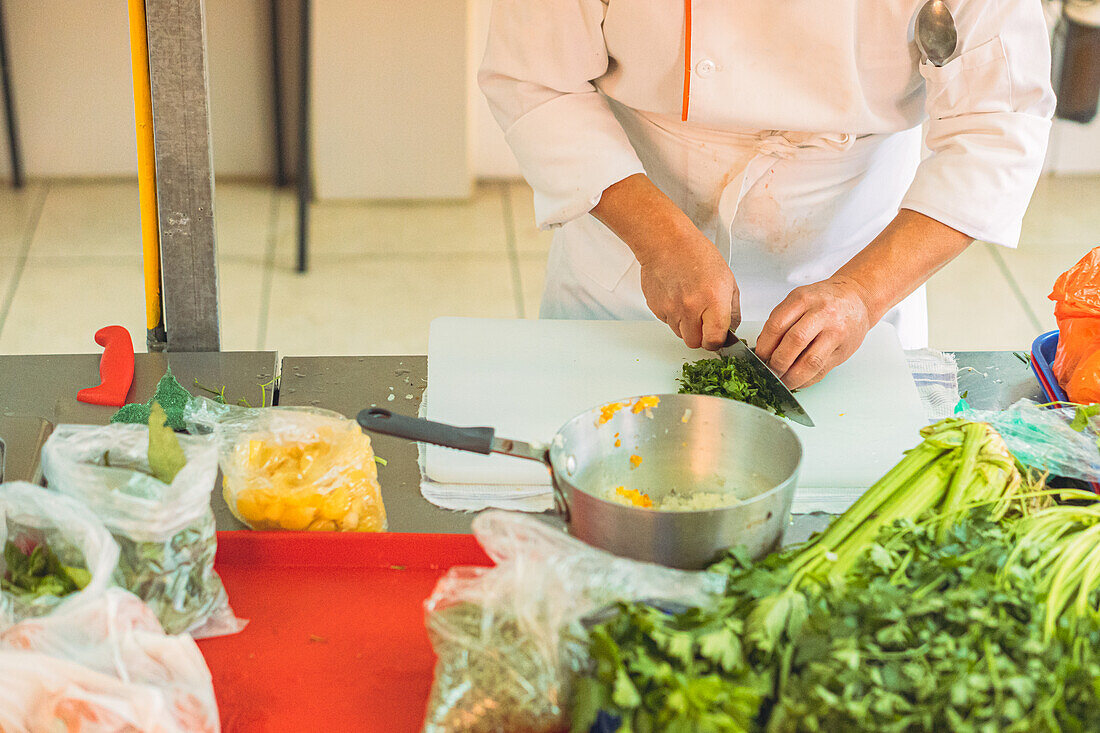 Anonymer Küchenchef in Uniform schneidet frische grüne Petersilie mit einem Messer auf einem Schneidebrett in der Küche