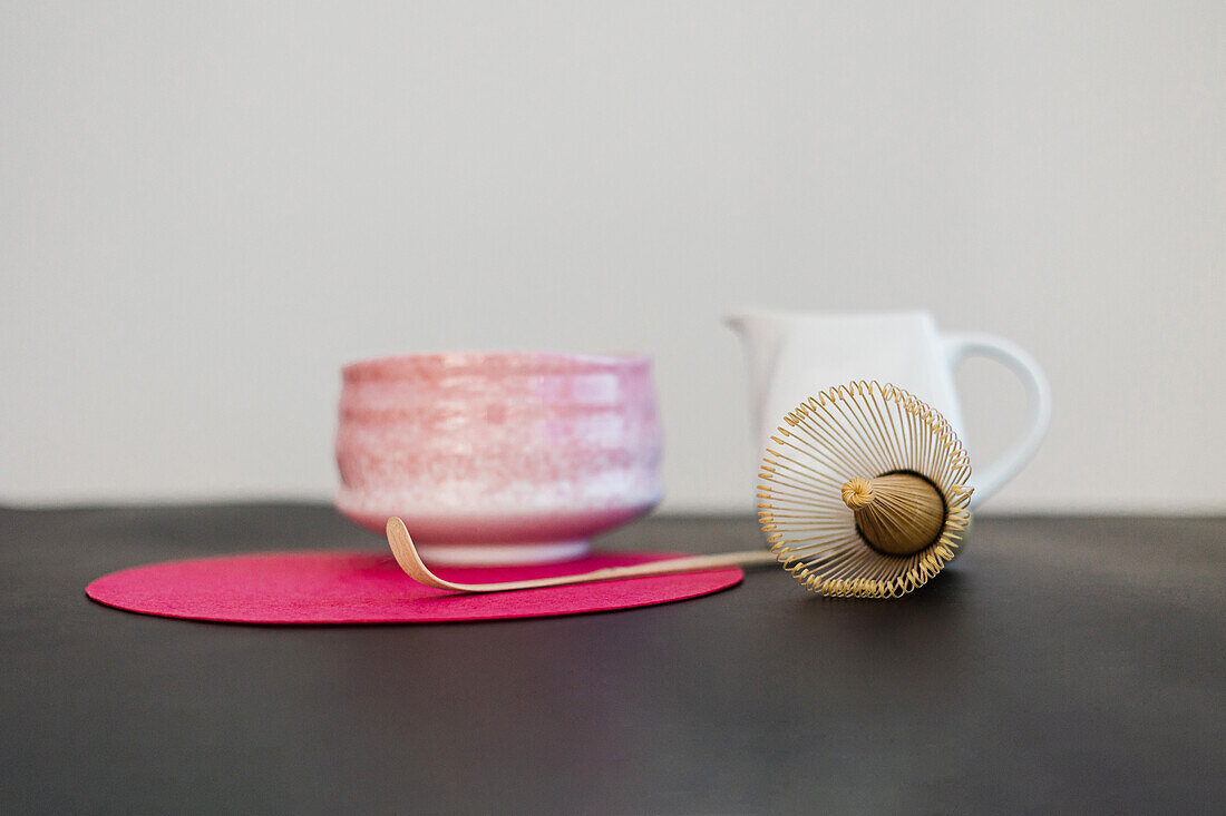 Keramikschüssel und hölzerne Chasen und Chashaku auf rosa rundem Ständer auf schwarzem Tisch im Sonnenlicht