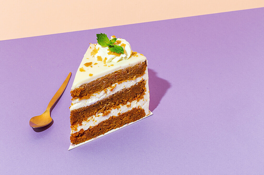 Slice of tasty sweet carrot sponge cake with cream decorated with mint leaf served on plate with spoon on table on colorful background