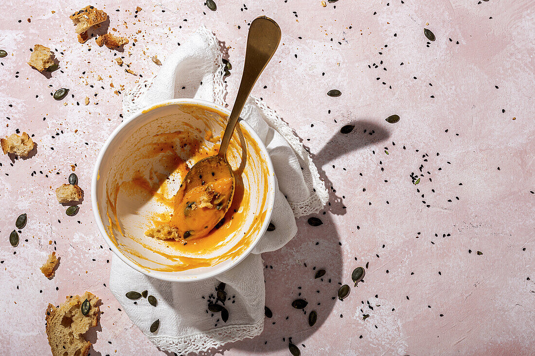 From above of spoon in white ceramic bowl with eaten pumpkin cream soup placed on table near scattered seeds and crispy bread pieces