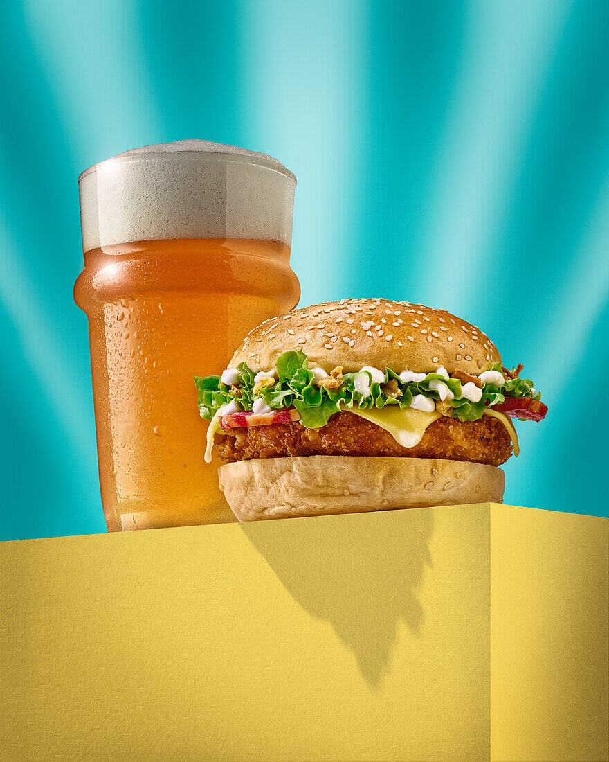 Leckerer Burger mit frischem Brötchen auf gelbem Block neben einem Glas mit kaltem, schäumendem Bier vor türkisem Hintergrund