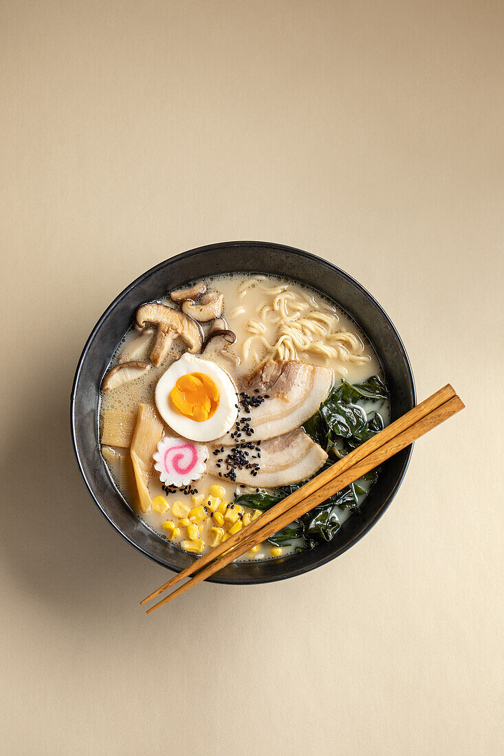 From above of appetizing Japanese ramen with boiled egg and mushrooms served in bowl with wooden chopsticks against beige background