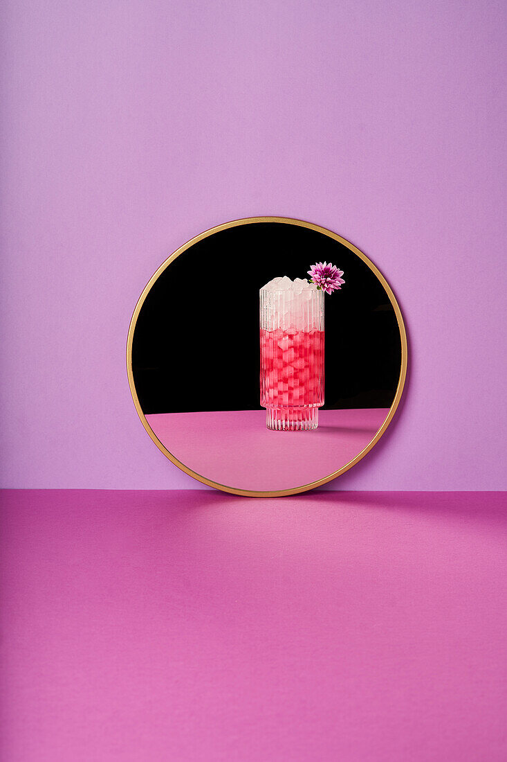 Glas mit erfrischendem Cocktail und Eiswürfeln, verziert mit einer Blume, auf einem hellvioletten Tisch vor einem Spiegel