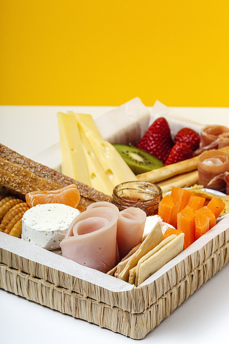 Brunchbox mit verschiedenen Wurst- und Käsesorten und Knäckebrot, arrangiert neben reifen Kiwi, süßen Erdbeeren und geschälten Mandarinen, neben Marmelade in einem Glas auf buntem Hintergrund