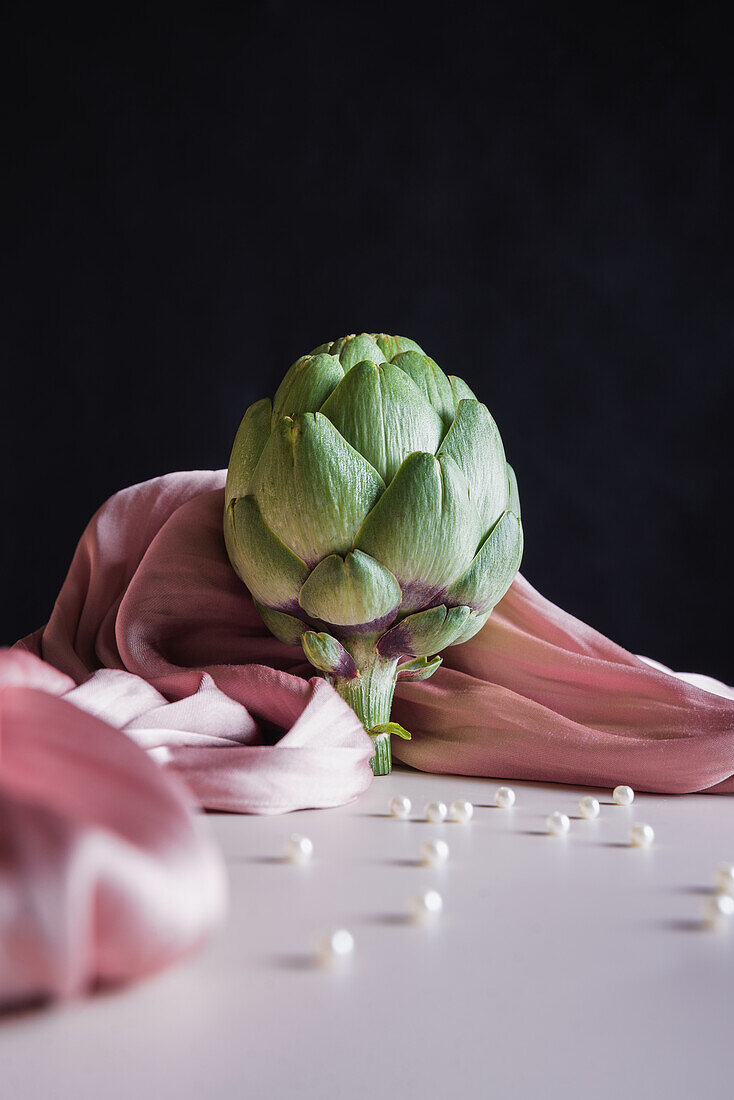 Composition of fresh healthy artichoke placed on draped fabric on white table with small beads and black ground