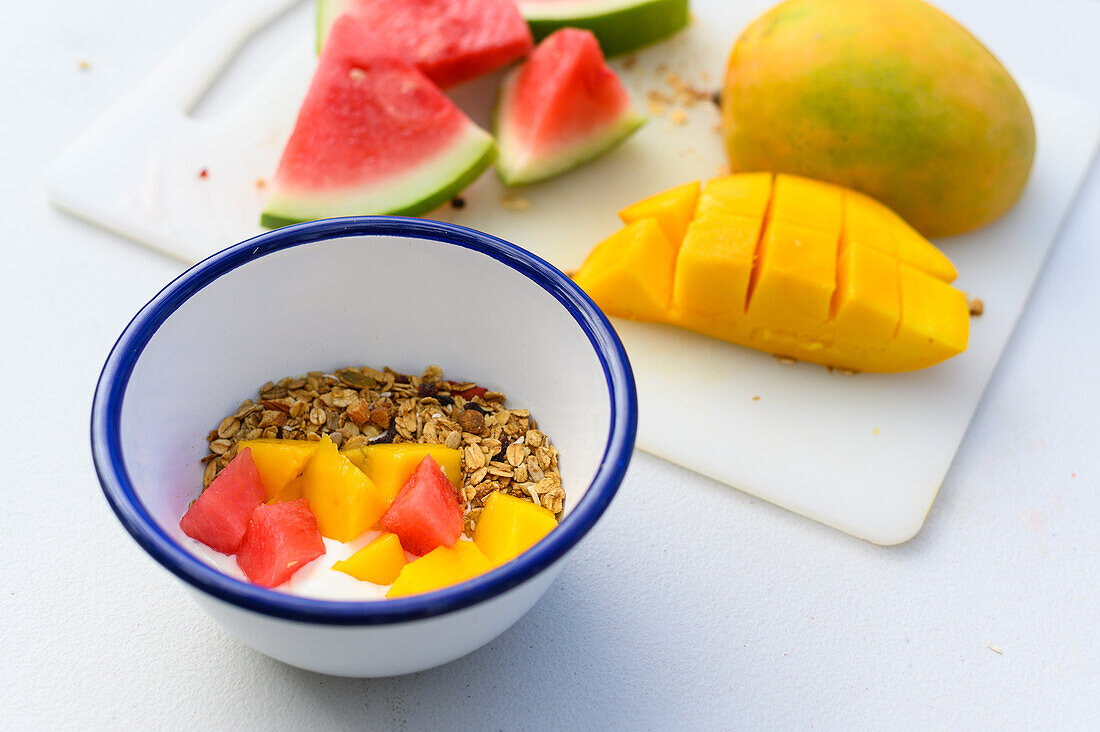 High angle of bowl with yogurt seeds slices of mango and watermelon placed on table near chopping board