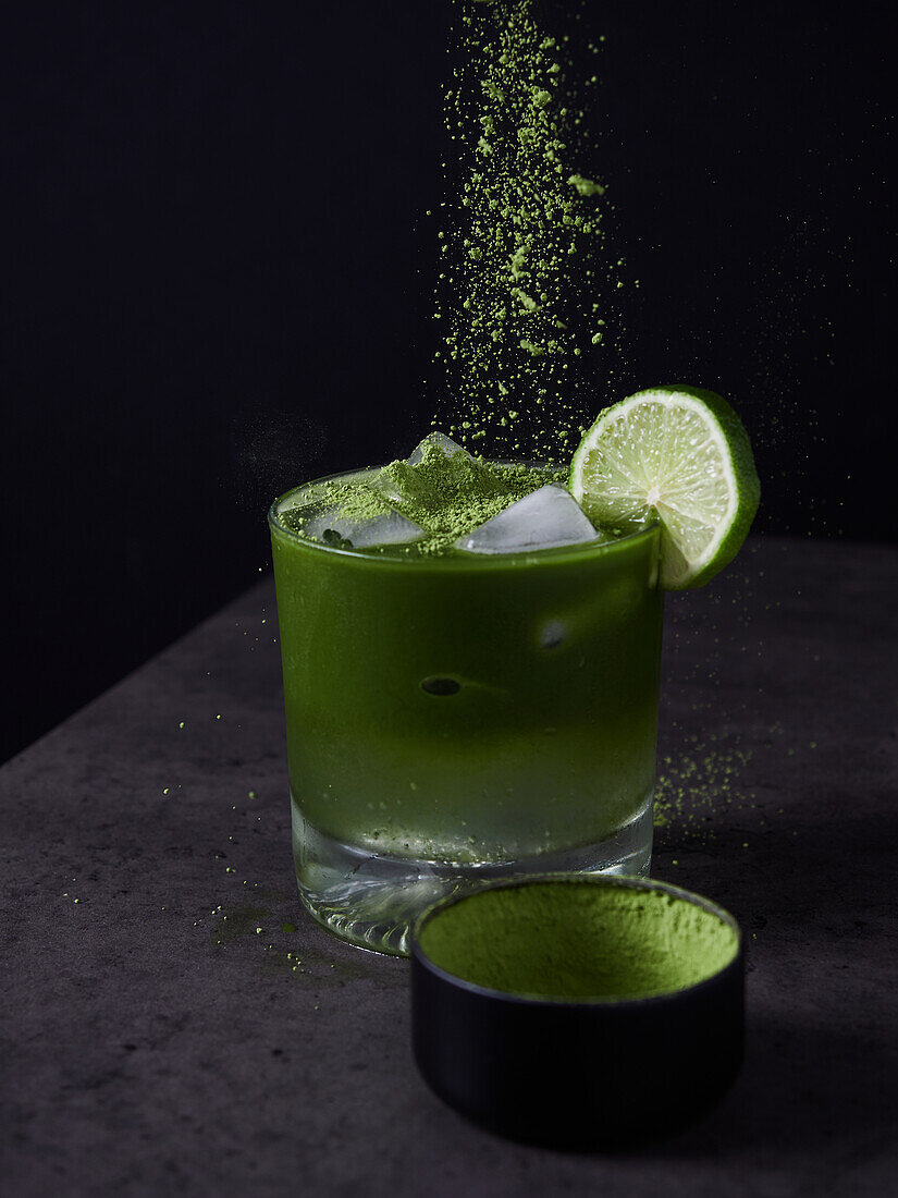 Refreshing cold drink with lime and matcha powder with ice cubes on dark surface