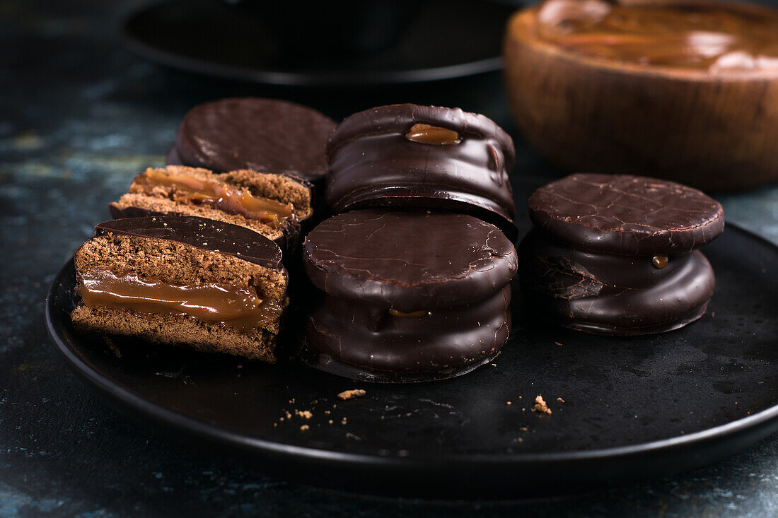 Schokoladen-Alfajores auf einem Teller und Dulce de Leche in einer Schüssel neben einer Tasse mit heißer Schokolade