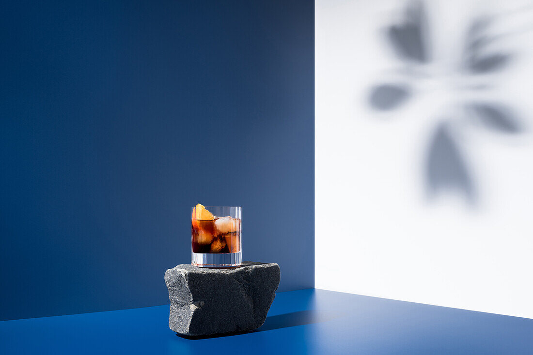 Glass of alcoholic cocktail made with red vermouth served with orange slices on stone in light studio with blue and white wall with shadow