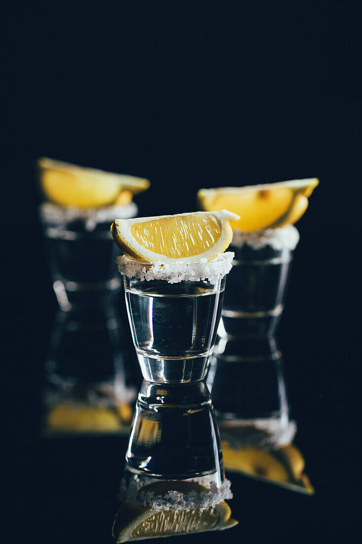 Tequila shots with salt and lemon placed on reflective surface against dark background