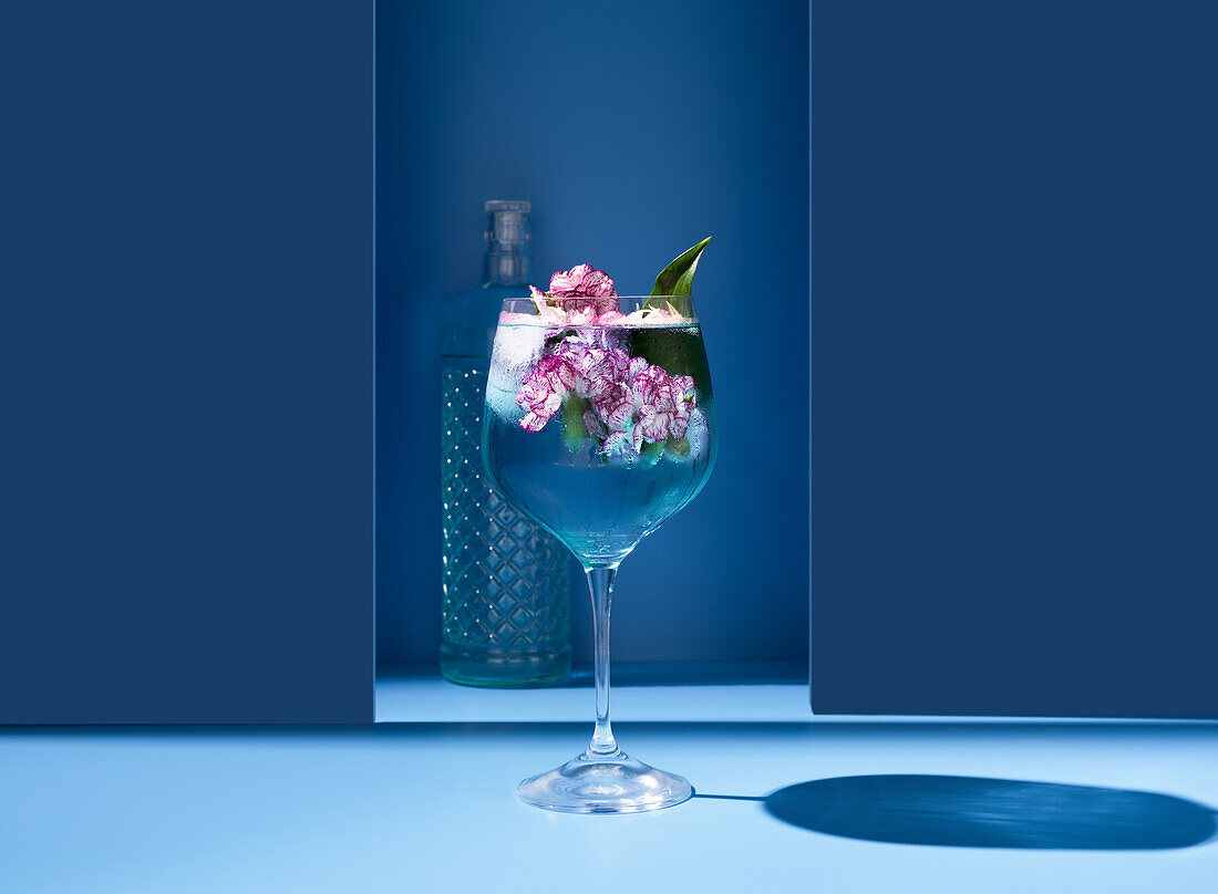 Transparent glass of fresh cocktail with mint leaves and flowers placed on surface near bottle against blue background