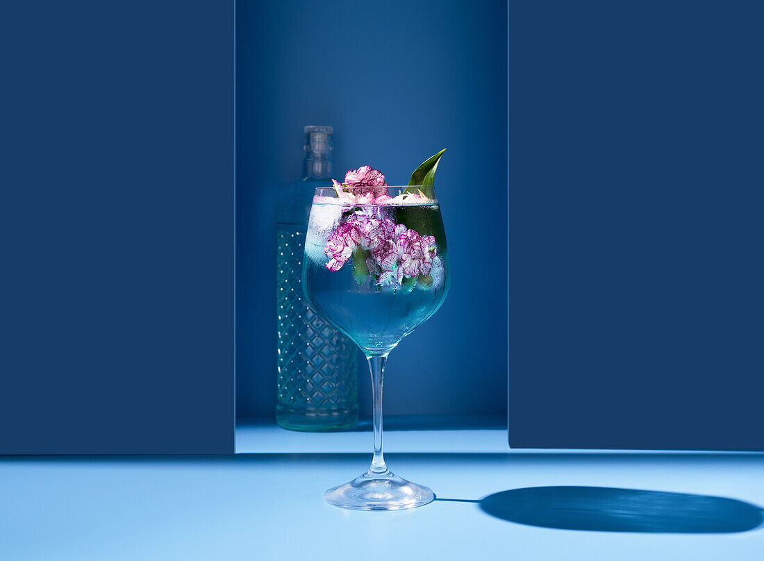 Transparentes Glas eines frischen Cocktails mit Minzblättern und Blumen auf der Oberfläche neben der Flasche vor blauem Hintergrund