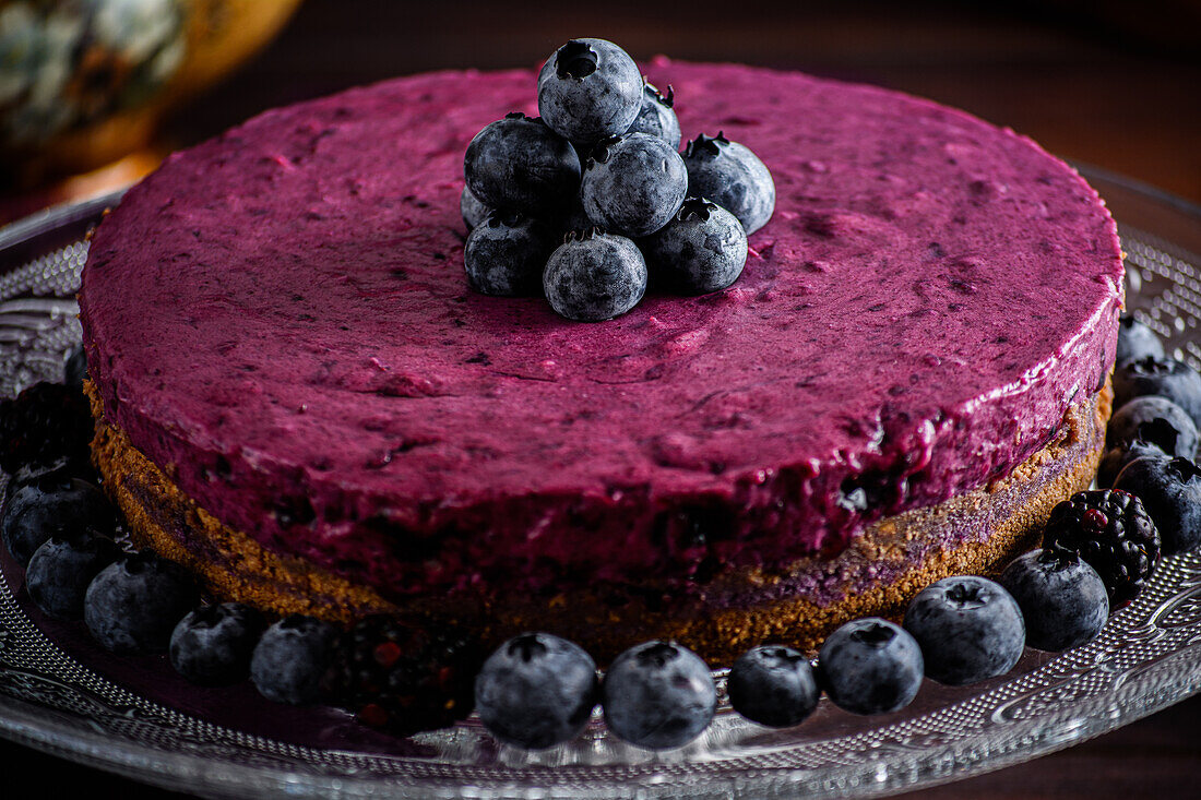 Delicious blueberry mousse cake with purple cream decorated with fresh berries served on glass stand on dark table