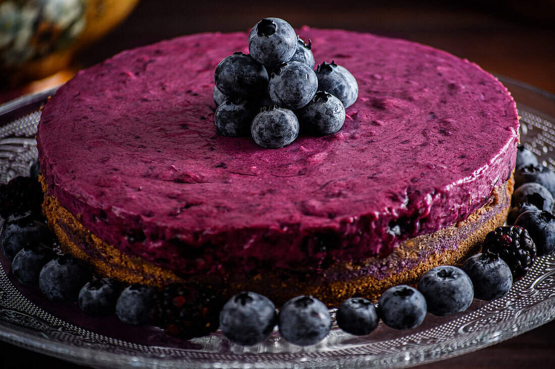 Köstliche Blaubeer-Mousse-Torte mit violetter Sahne, dekoriert mit frischen Beeren, serviert auf einem Glasständer auf einem dunklen Tisch