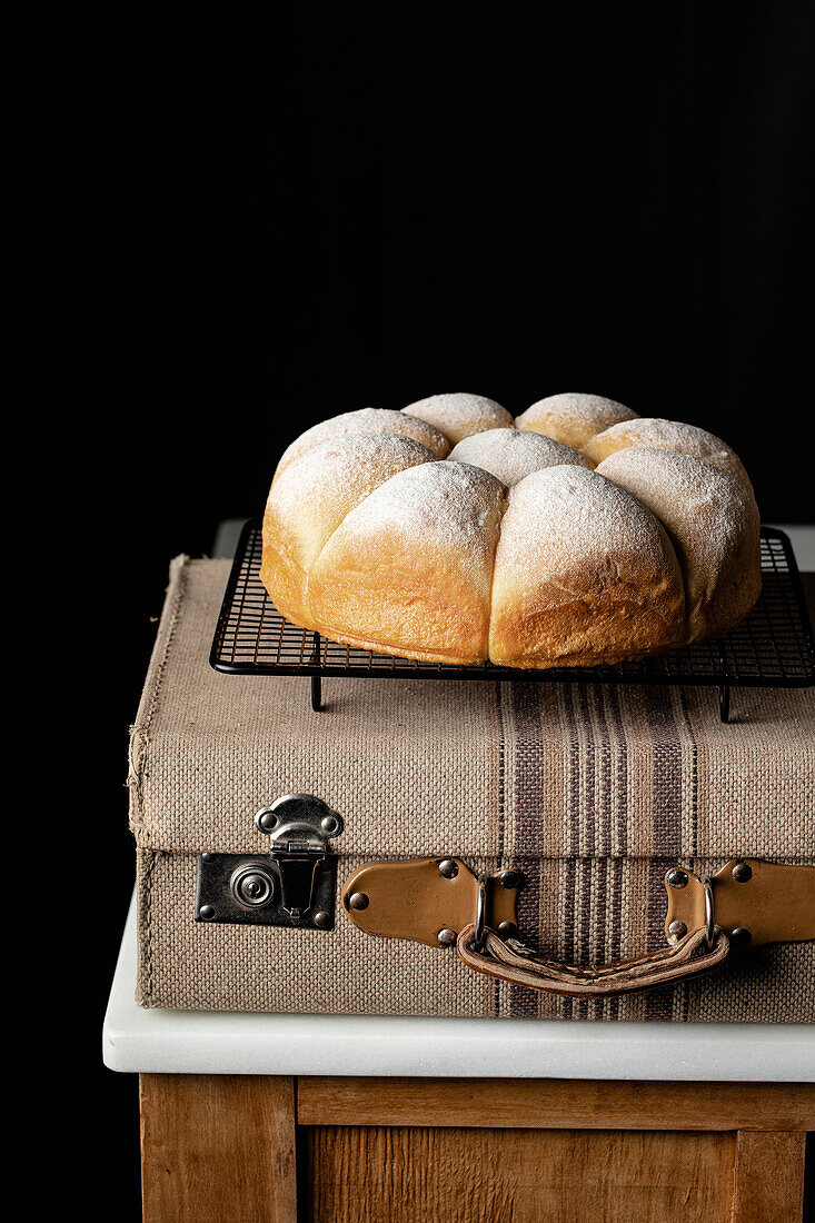 Appetizing soft bread buns on baking tray placed on suitcase near black wall