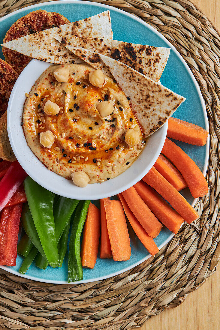 Schale mit Hummus und Tortilla-Chips, umgeben von verschiedenen Gemüsesorten und Croutons, serviert auf einer Weidenmatte auf einem Holztisch, von oben gesehen