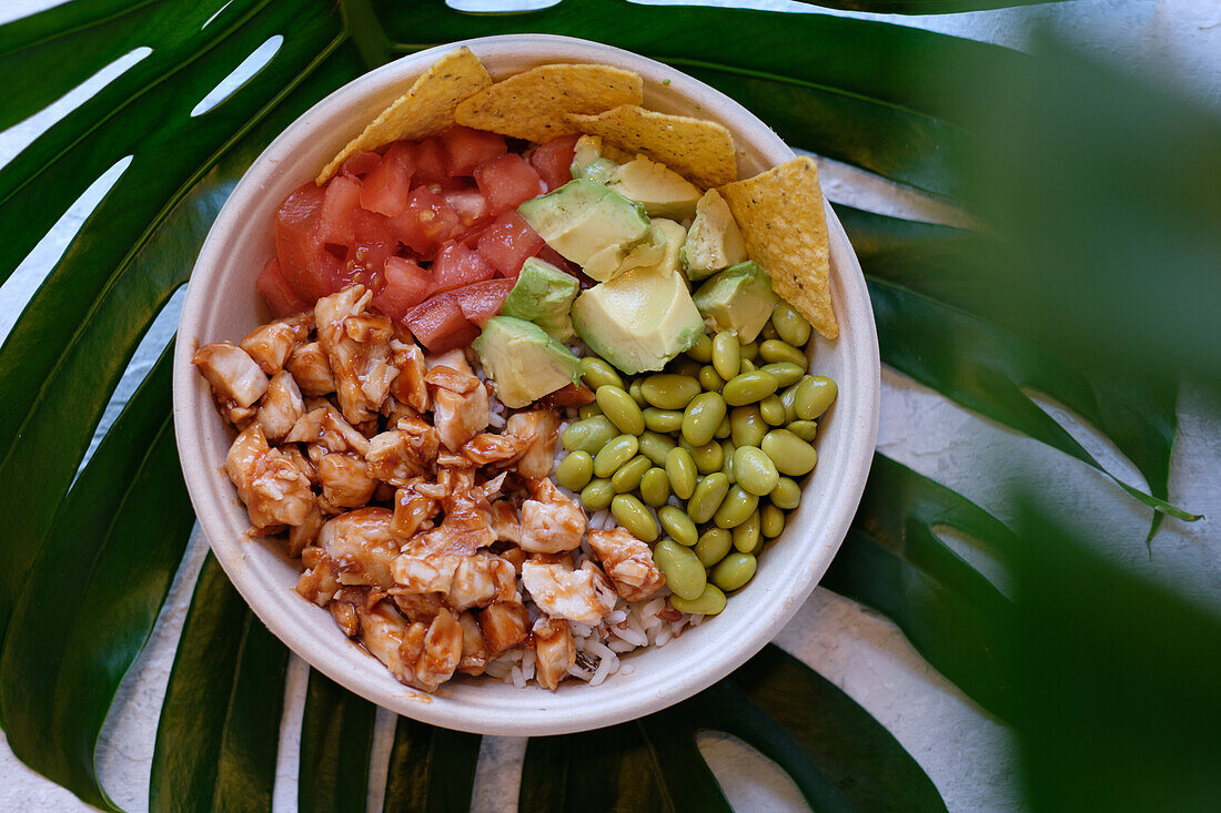 Top view of appetizing Hawaiian rice bowl with saucy chicken and avocado served with fresh tomato beans and nachos