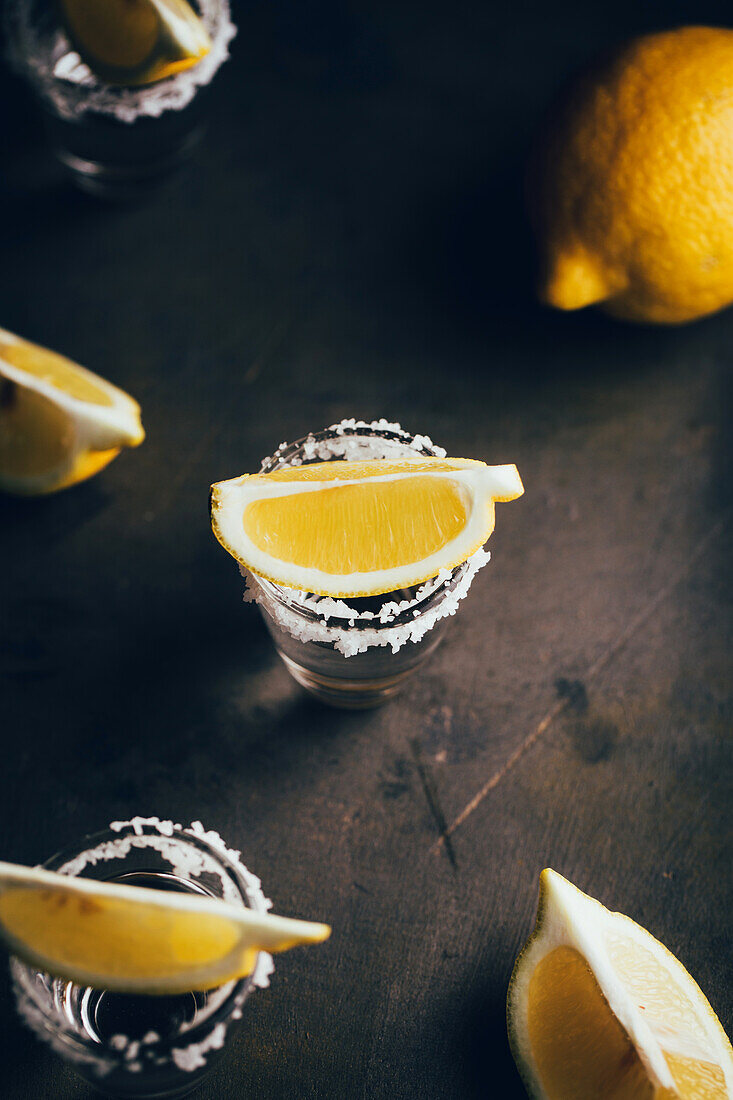 Top view of tequila shots with salt and lemon placed on rustic surface against dark background