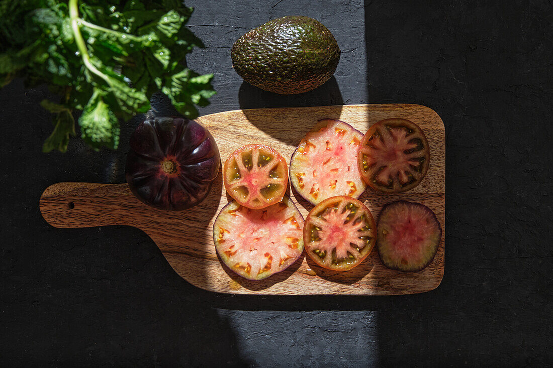 Top view of fresh ripe sliced black tomatoes and green mint stems on wooden cutting board on black background