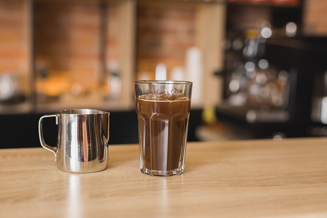 Glasbecher mit aromatischem, geeistem Americano auf einem Stehtisch neben einem Metallkrug in einem hellen Cafe