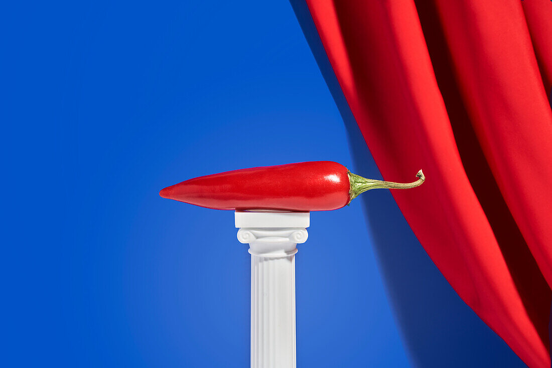 Red hot pepper placed on miniature column against blue background with red velvet curtain